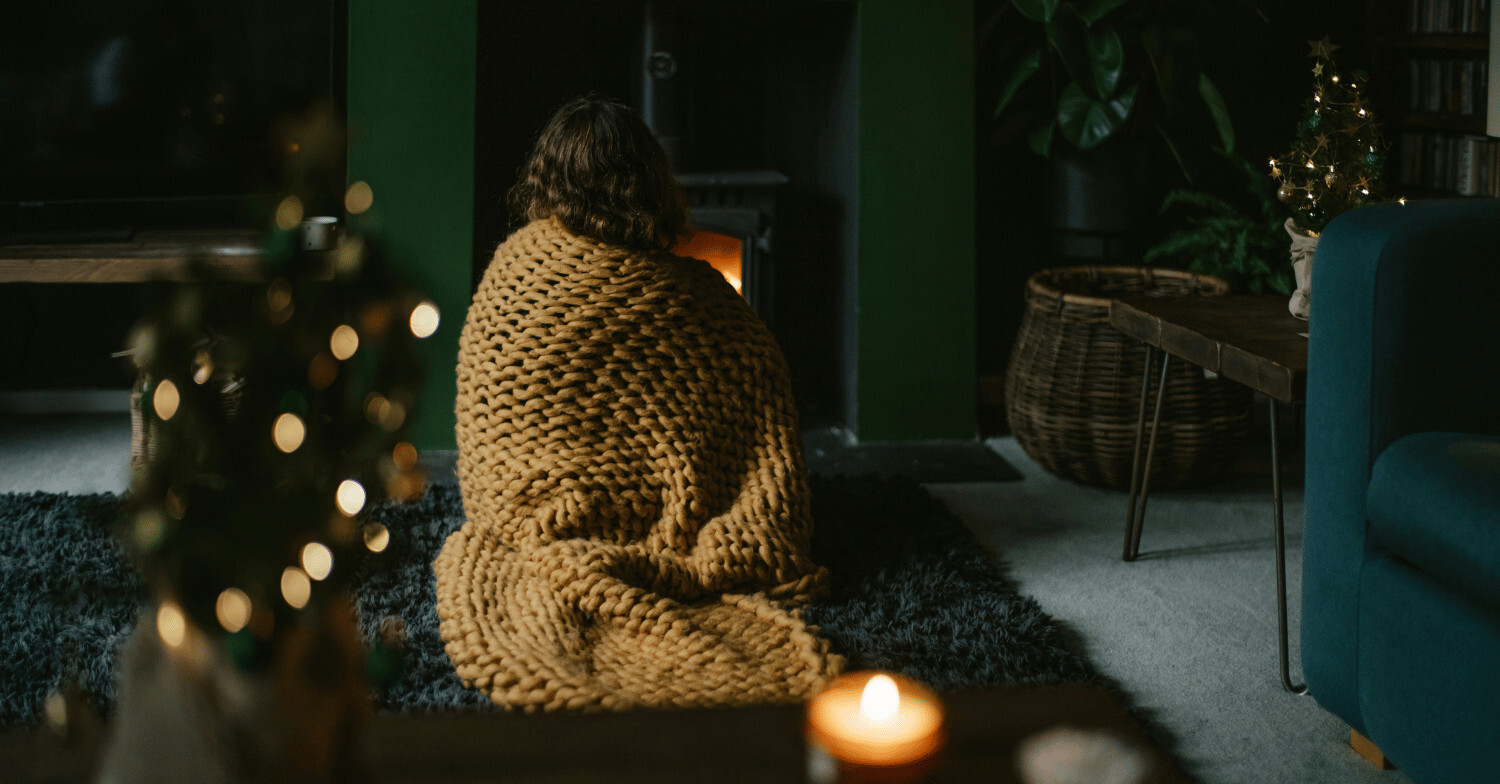 Person wrapped in a knitted blanket sitting alone near a fireplace with holiday lights, reflecting emotional exhaustion, quiet reflection, and the need for rest during the holiday season.